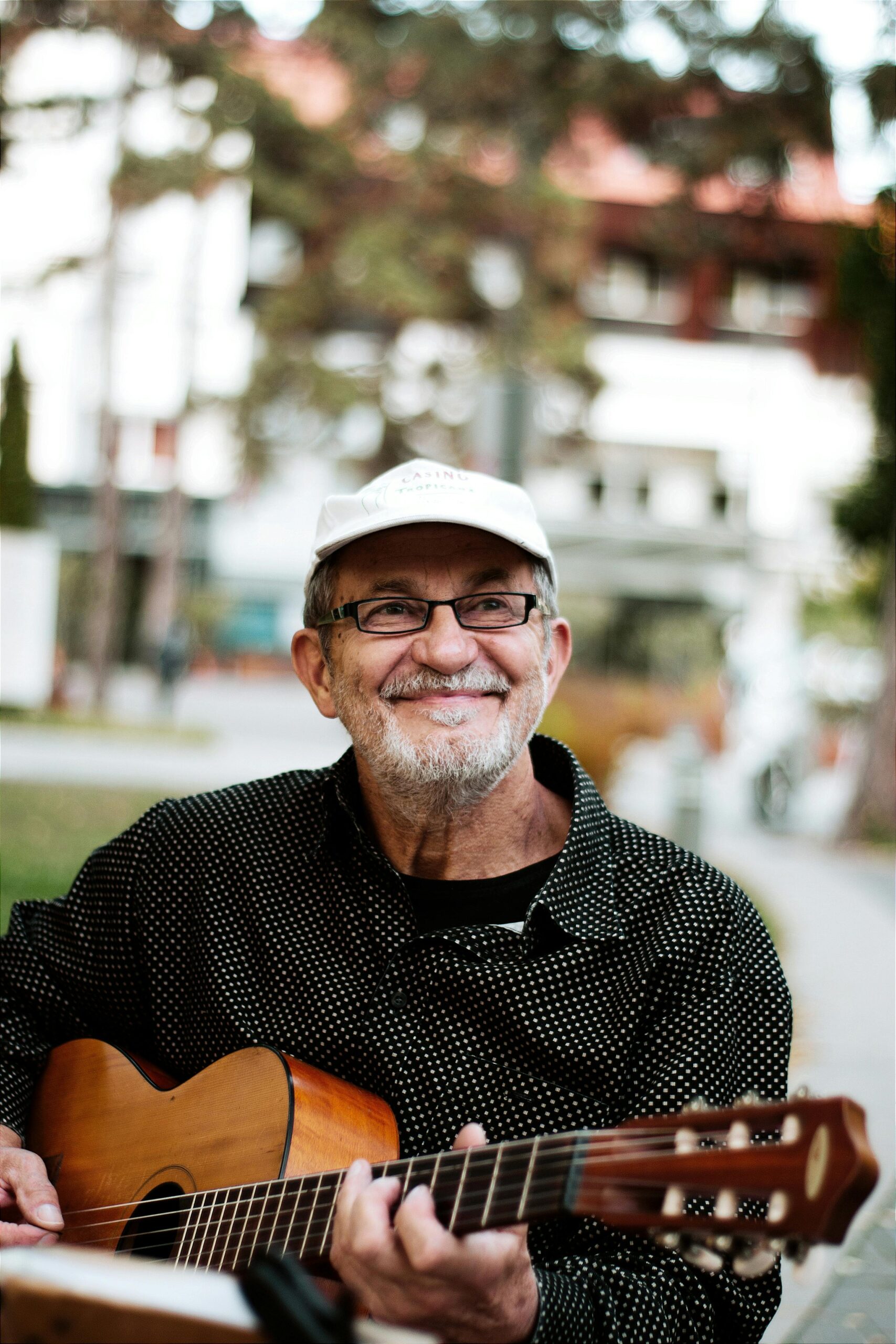 Elderly man with glasses and hat plays guitar outside, smiling happily.