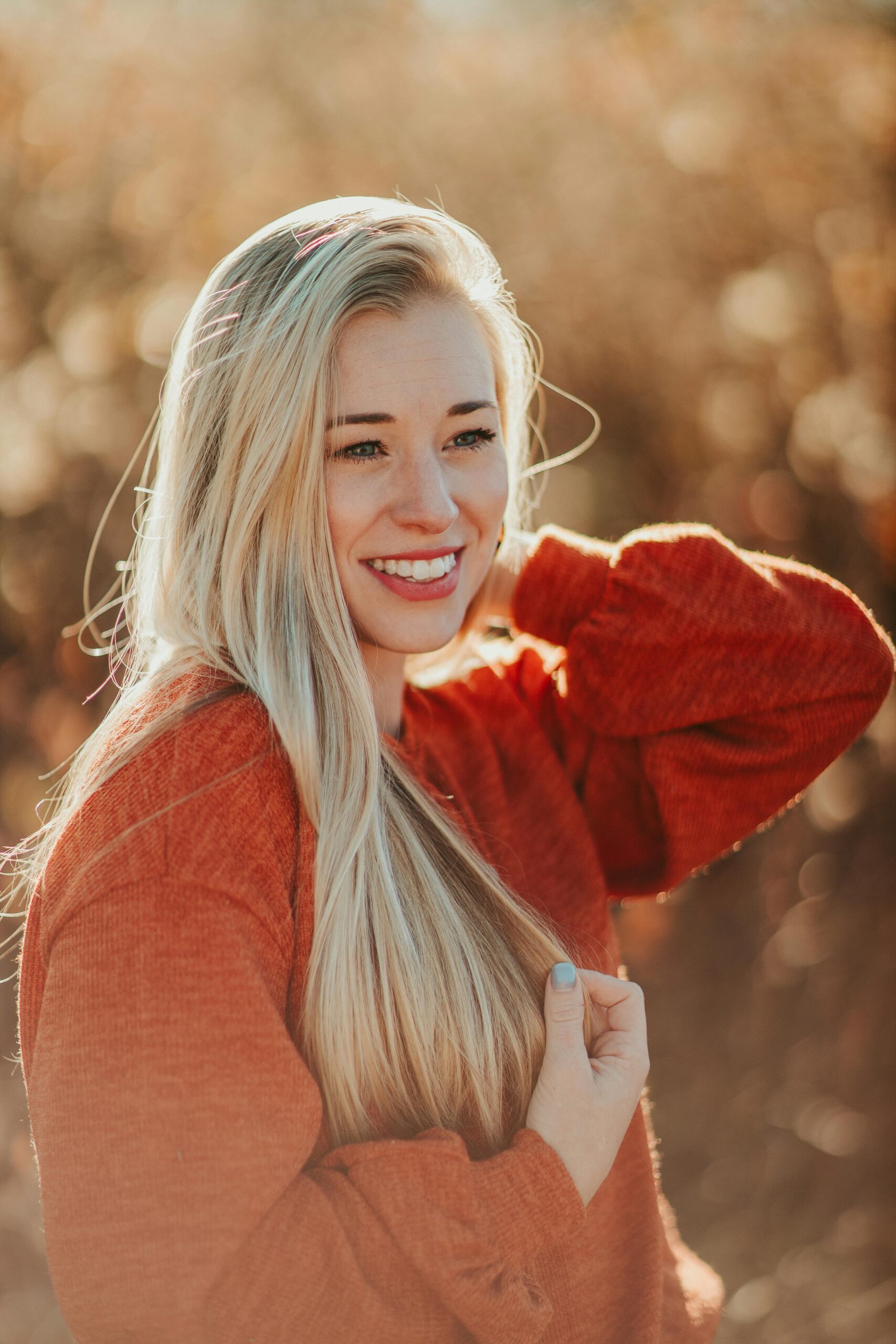 Attractive cheerful female wearing cozy red sweater touching hair tenderly and looking away happily while standing on blurred nature background on sunny autumn day